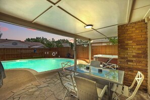 A covered patio with a glass table and chairs, a brick wall, and a backyard pool. - Modern & Chic 4-bd/2-ba Pool, Hot Tub, BBQ Grill (Carrollton)