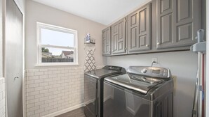 A laundry room with two stainless steel washers and dryers, a window with a view, and a tiled backsplash. - Modern & Chic 4-bd/2-ba Pool, Hot Tub, BBQ Grill (Carrollton)