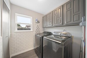 A laundry room with two stainless steel washers and dryers, a window with a view, and a tiled backsplash. - Modern & Chic 4-bd/2-ba Pool, Hot Tub, BBQ Grill (Carrollton)
