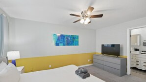 A hotel room with a bed, a ceiling fan, a TV, a dresser, and a painting on the wall. - Modern & Chic 4-bd/2-ba Pool, Hot Tub, BBQ Grill (Carrollton)