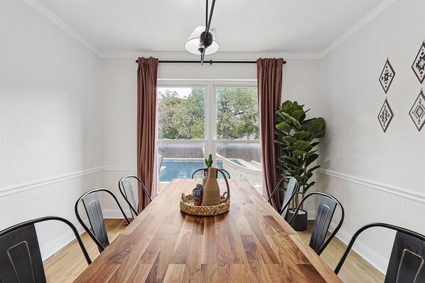 A dining area with a wooden table, metal chairs, a vase centerpiece, and a view of a pool through large windows. - 5BR w/ Backyard Paradise, Pool, Cinema, games (North Richland Hills)