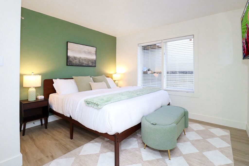 Une chambre à coucher avec un lit, des lampes de chevet, un mur vert, une fenêtre avec des stores et un tableau.