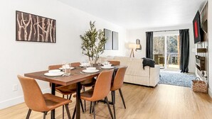 Uma sala de jantar com mesa de madeira posta para quatro pessoas, um sofá e uma televisão ao fundo.