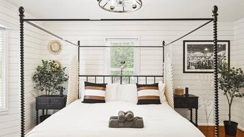 A four-poster bed with a canopy, a bedside table, a nightstand, a potted plant, and a framed picture on the wall.