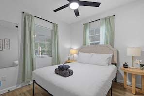 A hotel room with a bed, a ceiling fan, a nightstand with a lamp, and a window with curtains. - Inactive (Sherman)