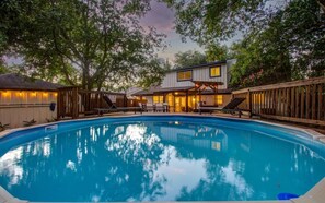 Ein Schwimmbecken mit Holzterrasse und Liegestühlen, umgeben von Bäumen und einem Haus mit überdachter Terrasse.