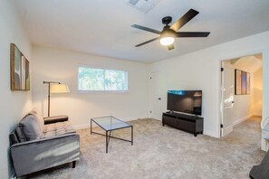 A living room with a sofa, a glass coffee table, a TV, and a ceiling fan. - Stunning Modern Dream Home w Pool, Grill, Foosball (Houston)