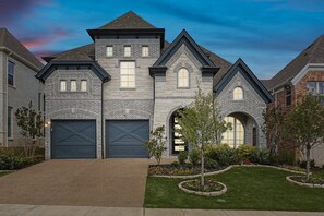 A two-story house with a garage, a well-maintained lawn, and a clear sky. - Brand New 5bd, 4BA Luxury Vacation Home w Hot Tub (Irving)