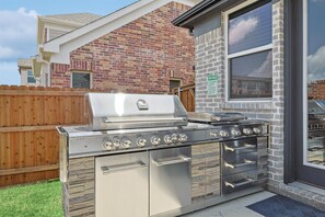A stainless steel outdoor kitchen with a grill, oven, and drawers, adjacent to a brick building with a window. - Brand New 5bd, 4BA Luxury Vacation Home w Hot Tub (Irving)
