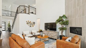 A spacious living area with a staircase, leather sofas, a flat-screen TV, and a potted plant. - Brand New 5bd, 4BA Luxury Vacation Home w Hot Tub (Irving)