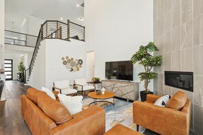 A spacious living area with a staircase, leather sofas, a flat-screen TV, and a potted plant. - Brand New 5bd, 4BA Luxury Vacation Home w Hot Tub (Irving)