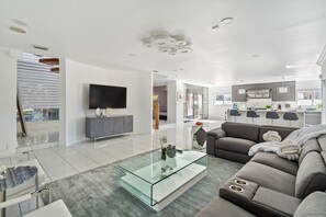 A modern living room with a glass coffee table, a grey sofa, and a flat-screen TV. - Tropical Pool Paradise x 6-bd, 4-ba Luxury Living (Miami Lakes)