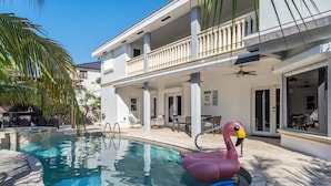 A swimming pool with a pink flamingo float, a covered outdoor seating area, and a two-story building with balconies. - Tropical Pool Paradise x 6-bd, 4-ba Luxury Living (Miami Lakes)