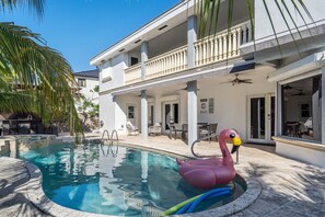 A swimming pool with a pink flamingo float, a covered outdoor seating area, and a two-story building with balconies. - Tropical Pool Paradise x 6-bd, 4-ba Luxury Living (Miami Lakes)