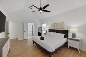 Une chambre à coucher comprenant un lit, des tables de chevet, un ventilateur de plafond et une porte donnant sur une autre pièce.