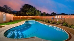 A swimming pool with a wooden deck, a hammock, and a fence with string lights. - Boho Chic Home w Backyard Paradise - Pool + Grill (North Richland Hills)