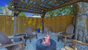 Un espacio de estar al aire libre techado, con sillas Adirondack, un pozo de fuego y una pérgola de madera.