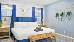 Habitación de hotel con cama, mesita de noche de madera, lámpara de noche y una planta en la pared, además de una alfombra estampada.