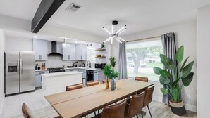 A modern kitchen with white cabinets, a stainless steel refrigerator, a dining table with chairs, and a large potted plant. - Relaxing Texas Retreat, 4BR w Pool & Dream Kitchen (DeSoto)