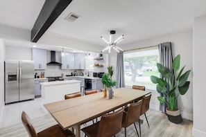 A modern kitchen with white cabinets, a stainless steel refrigerator, a dining table with chairs, and a large potted plant. - Relaxing Texas Retreat, 4BR w Pool & Dream Kitchen (DeSoto)