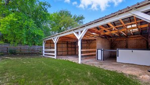A covered outdoor area with a wooden structure, a fenced yard, and a large white building in the background. - 5-bd/2.5-ba Modern Farmhouse Retreat (Flower Mound)
