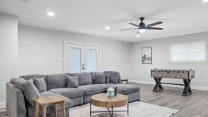 A living room with a gray sectional sofa, a round wooden coffee table, a foosball table, and a ceiling fan. - 5-bd/2.5-ba Modern Farmhouse Retreat (Flower Mound)