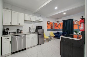 Eine kompakte Küche mit weißen Schränken, einem Edelstahlkühlschrank, einer Mikrowelle und einem Spülbecken. Es gibt einen Essbereich mit einem blauen Sofa und gelben Stühlen.