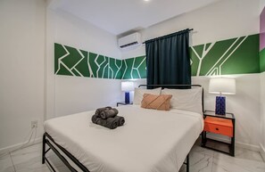 A hotel room with a bed, two bedside tables, a wall-mounted air conditioner, and a geometric-patterned wall. - Charming Houston Getaway 2bd/1ba & yard (Houston)