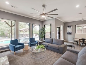 A spacious living room with a large ceiling fan, a gray sofa, two blue armchairs, a glass coffee table, and a view of a backyard with a pool. - Clearview Castle - Pool, Jacuzzi, Lake View (Lewisville)