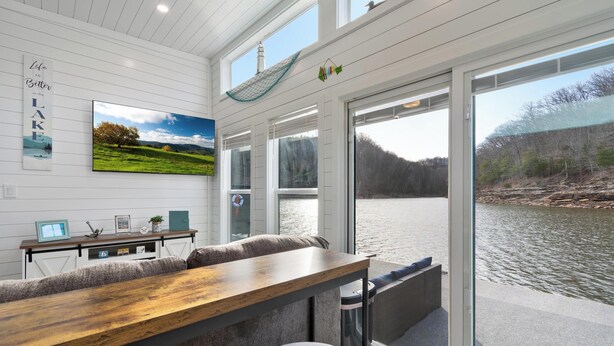 A modern interior with a wooden table, cushioned seating, a large window overlooking a lake, and a framed landscape painting. - Dock Holiday - Unique Floating House on Cumberland (Russell Springs)