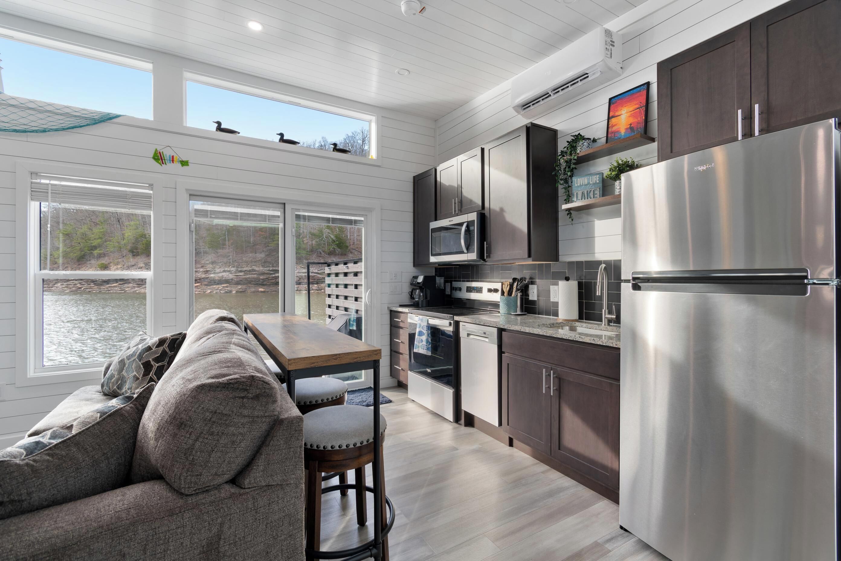 A modern kitchen with a stainless steel refrigerator, dark wooden cabinets, and a view of a lake through large windows.