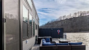 A waterfront patio with outdoor seating, a large window, and a view of the water and trees. - Dock Holiday - Unique Floating House on Cumberland (Russell Springs)