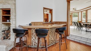 Uma área de bar com balcão de madeira, três banquetas pretas, um espelho e uma parede de pedra.
