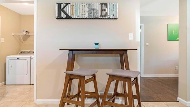 A kitchen area with a 'Kitchen' sign, two wooden stools, a small table, and a washing machine in the background. - Spacious Luxurious House, Big Groups and Pool (Irving)