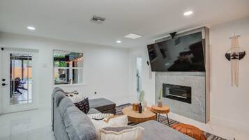 A modern living room with a grey sofa, a flat-screen TV mounted above a fireplace, and a large window with a view of the outdoors.