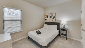 A bedroom with a bed, a bedside table with a lamp, a window with blinds, and two wall art pieces. - Your Vibrant Dallas Getaway Home (Dallas)