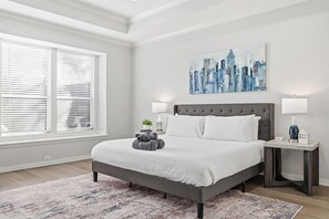 A bedroom with a large bed, two bedside tables, a cityscape painting, and a window with blinds. - Luxury Backyard Pool & Patio Oasis (Lewisville)