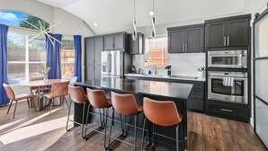 A modern kitchen with dark cabinets, stainless steel appliances, a large island with bar stools, and a dining area with a table and chairs. - 5-bd/2-ba Vacation Retreat w/ Pool, Hot Tub & bbq (Fort Worth)