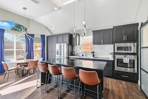A modern kitchen with dark cabinets, stainless steel appliances, a central island with bar stools, and a dining area with a table and chairs. - 5-bd/2-ba Vacation Retreat w/ Pool, Hot Tub & bbq (Fort Worth)