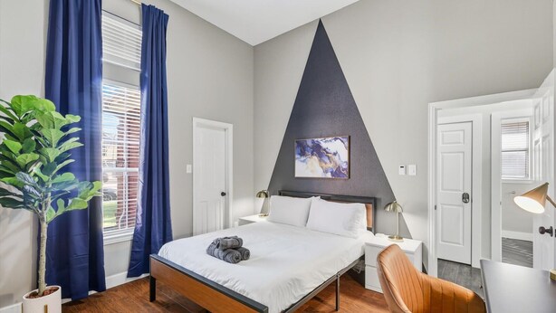 A modern bedroom with a bed, bedside tables, a chair, a potted plant, and a painting on the wall. - 5-bd/2-ba Vacation Retreat w/ Pool, Hot Tub & bbq (Fort Worth)