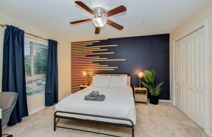 Quarto com cama, ventoinha no teto, janela com cortinas, mesa de cabeceira com abajur e planta em vaso.