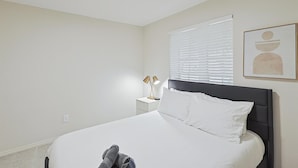 Ett sovrum med en säng, ett sängbord med en lampa, ett fönster med persienner och ett konstverk som hänger på väggen.