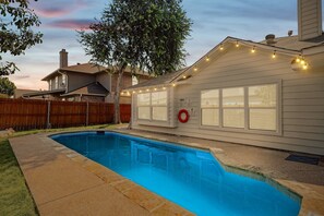 A residential swimming pool with a wooden deck, a wooden fence, and a house with large windows and string lights. - 4bd, 2BA Saginaw Sanctuary - Pool, Firepit, Laundry (Fort Worth)