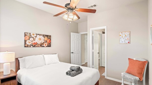 Un dormitorio con cama, mesita de noche, una silla, ventilador de techo y una puerta.