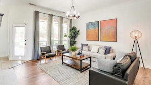 A modern living room with a sofa, two armchairs, a coffee table, and a floor lamp. There are two paintings on the wall and a potted plant on the coffee table. - Southern Charm & Luxury Living - BBQ & Pool (Rockwall)