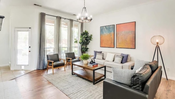 A modern living room with a sofa, two armchairs, a coffee table, and a floor lamp. There are two paintings on the wall and a potted plant on the coffee table. - Southern Charm & Luxury Living - BBQ & Pool (Rockwall)