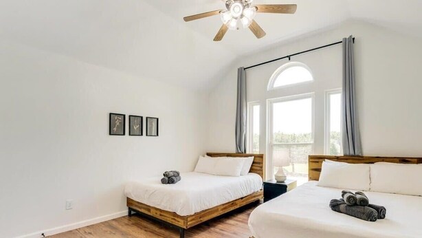 A bedroom with two wooden beds, a ceiling fan, a large window with curtains, and a wall-mounted mirror. - Southern Charm & Luxury Living - BBQ & Pool (Rockwall)