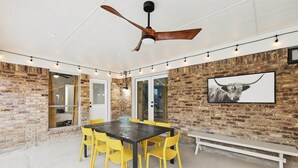 A covered patio area with a brick wall, a ceiling fan, a dining table with yellow chairs, and a bench. - Best of Desoto 4br,3ba With Pool, Hottub, Cinema (DeSoto)