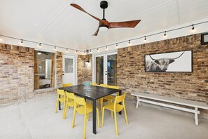 A covered patio area with a brick wall, a ceiling fan, a dining table with yellow chairs, and a bench. - Best of Desoto 4br,3ba With Pool, Hottub, Cinema (DeSoto)