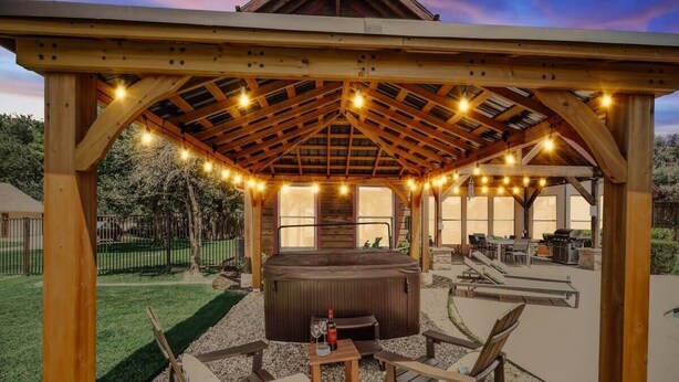 A wooden gazebo with string lights, a hot tub, and outdoor seating. - Rustic Cabin Mansion - Hot Tub, Gym, Cinema, Pool (McKinney)